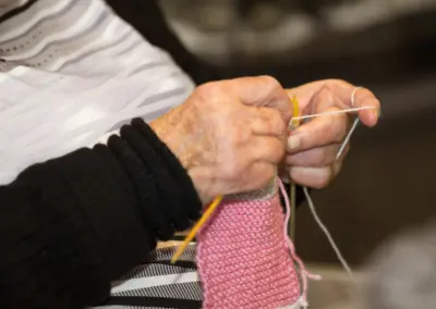 Nahaufnahme von Händen beim Stricken: rosa Garn, gelbe Stricknadel, pinkes Gestrick vor dunklem Hintergrund.