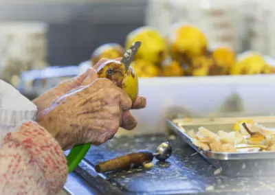 Hand mit Gummi-Handschuhen hält saftiges Brathähnchen am Grill, Küchenumgebung mit Beilagen im Hintergrund.