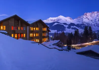 Winterliches Chaletdorf in den Alpen bei Dämmerung mit warm beleuchteten Fenstern, verschneiter Landschaft und Berggipfeln.