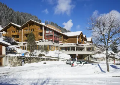 Moderne Holz-Chalets in winterlicher Berglandschaft mit Schnee, blauem Himmel, verschneitem Wald und Autos vor dem Gebäude.