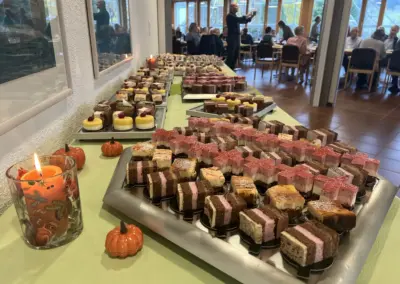 Vielfältige Desserts-Tafel mit Kuchen, Torten und Pralinen; kleine Kürbisse als Deko, Gäste genießen Speisen im Hintergrund.