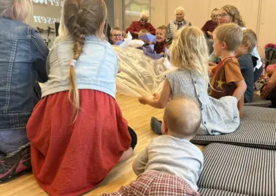 Kinder und ältere Menschen sitzen um einen Tisch und basteln Origami-Papierboote im Gemeinschaftsraum.