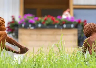 Bronzene Kinderskulpturen im Garten; Gras im Vordergrund, farbenfrohe Blumen im Hintergrund.