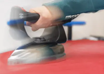 Hand mit türkisfarbener Schutzhandschuh bedient Exzenterschleifer auf rotem Untergrund.
