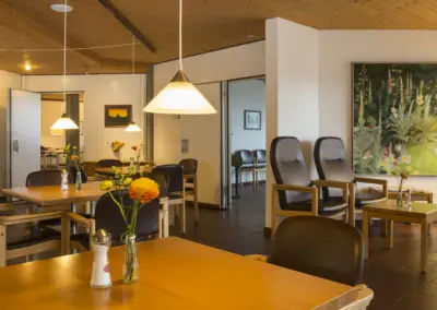 Gemütliches Café‑Interieur mit Holzboden, Holzdecke, bequemen Sesseln, Tischen, Pflanzen und warmem Licht.