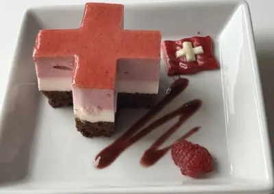 Mehrschichtiger Dessertkuchen in roter Kreuzform, rosa-weiße Schichten, Brownie-Boden, rote Sauce und Himbeeren.