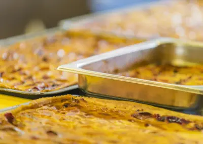 Buffet mit goldbraunen Aufläufen in Edelstahl-Backformen, Nahaufnahme einer Lasagne-ähnlichen Speise.