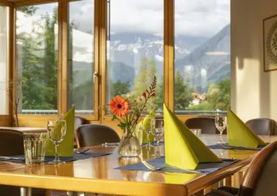 Gemütliches Restaurant mit großen Fenstern und Bergblick, gedeckter Tisch, gelbe Servietten und Blumenstrauß.