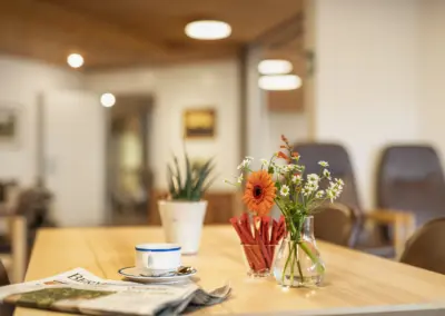 Blumenstrauß mit Gerbera auf Holztisch in modernem Café, Kaffeetasse und Zeitung im Vordergrund.