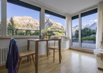 Helles Wohnzimmer mit großen Fensterfronten und Balkon; Blick auf Alpen, grüne Wiesen und Berge.