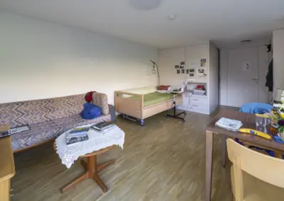 Helles WG-Zimmer mit Sofa, Einzelbett, Schreibtisch, Kleiderschrank und Tisch mit Blumen.