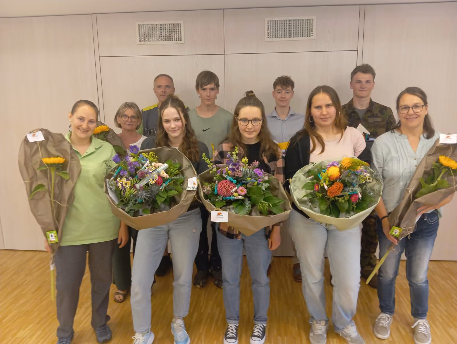 Gruppe junger Menschen mit bunten Blumensträußen posiert in einem Innenraum, vermutlich Abschlussfeier.